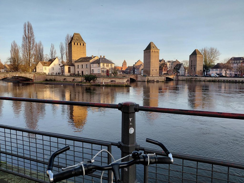 A bicycle locked to a railing on a bridge, with the medieval towers of the Ponts Couverts reflected in the River Ill in Strasbourg, France