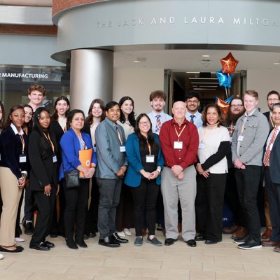 A diverse group of approximately 20 people wearing conference lanyards pose together for a group photo outside the Jack and Laura Milton Atrium, with red, blue, and orange star-shaped balloons visible in the background.