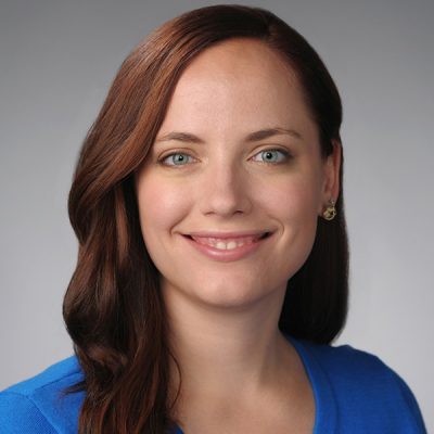 A woman with long auburn hair and blue eyes smiles in a professional headshot, wearing a blue top against a neutral gray background.