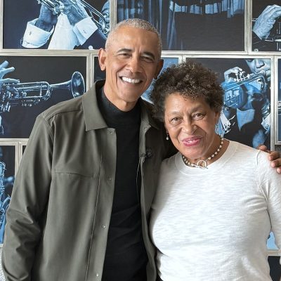 Two people smiling and posing together in front of a wall displaying black-and-white jazz photography prints.