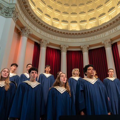The Hendricks Chapel Choir, in navy robes, performs beneath a domed rotunda with white columns, red curtains, and the inscription "They that worship Him must worship Him in Spirit."
