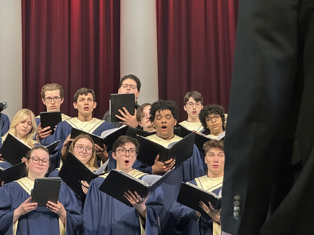 A youth choir in navy and gold robes sings from black folders before red curtains, with a conductor's silhouette visible in the foreground.