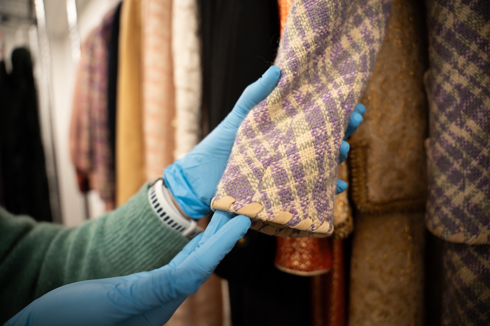 A gloved hand carefully handles a purple and cream plaid textile piece with a whipstitched leather edge, selecting it from a rack of stored garments and fabrics