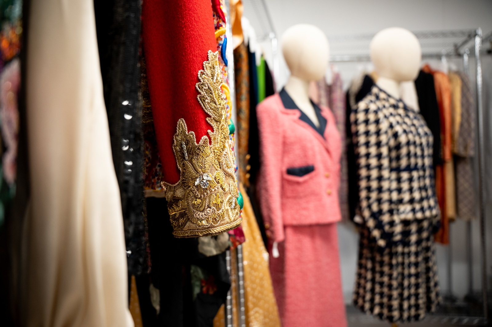 Garments displayed in a fashion collection storage area, with a red piece featuring ornate gold embroidery and jeweled embellishment in the foreground. Behind it, dress forms showcase a pink tweed skirt suit and a black-and-white houndstooth jacket, while additional garments hang on metal racks.