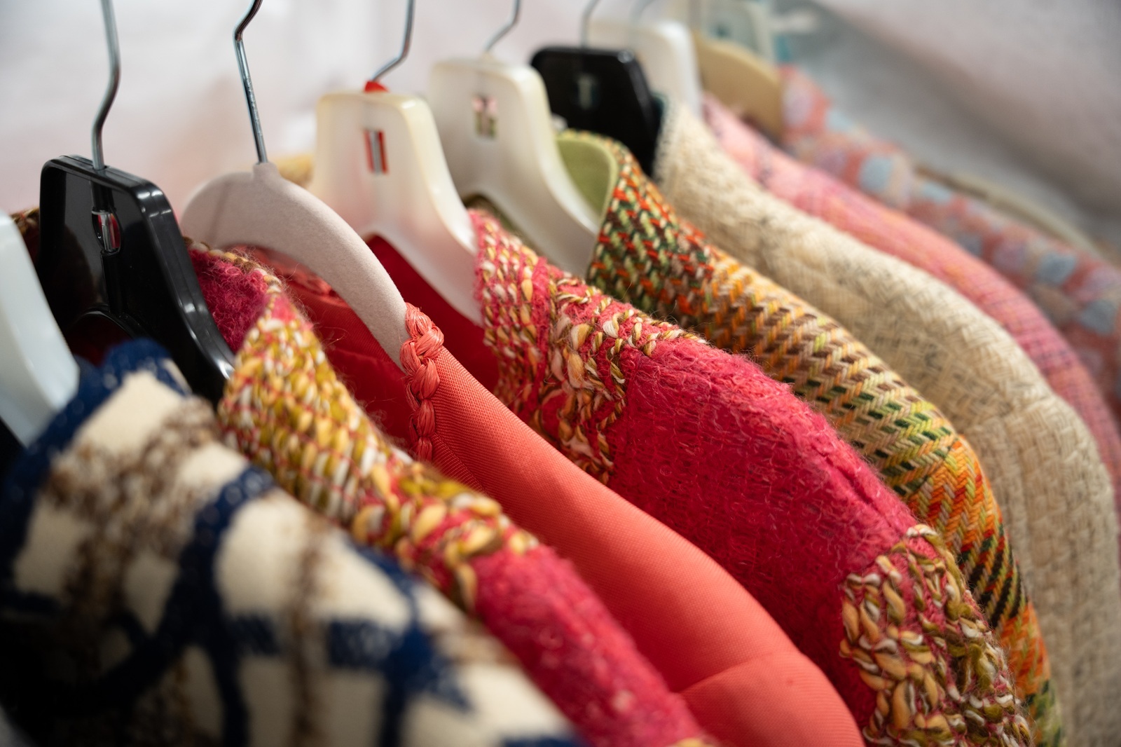 A close-up view of colorful garments hanging on a rack, showcasing a range of textured woven fabrics in vibrant pinks, reds, multicolored tweeds, and cream tones