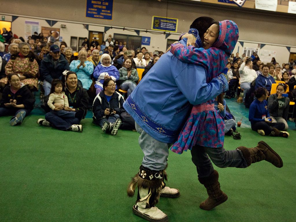 Two people in traditional Alaska Native clothing share a joyful embrace on the floor of a packed gymnasium, as a large crowd of smiling, applauding community members looks on