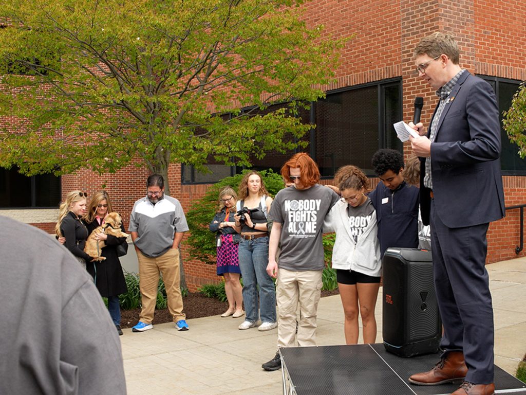 A man in a suit speaks into a microphone on a small stage at an outdoor event, while a group of attendees stands nearby with heads bowed. Several people wear "Nobody Fights Alone" shirts. One attendee holds a small dog.