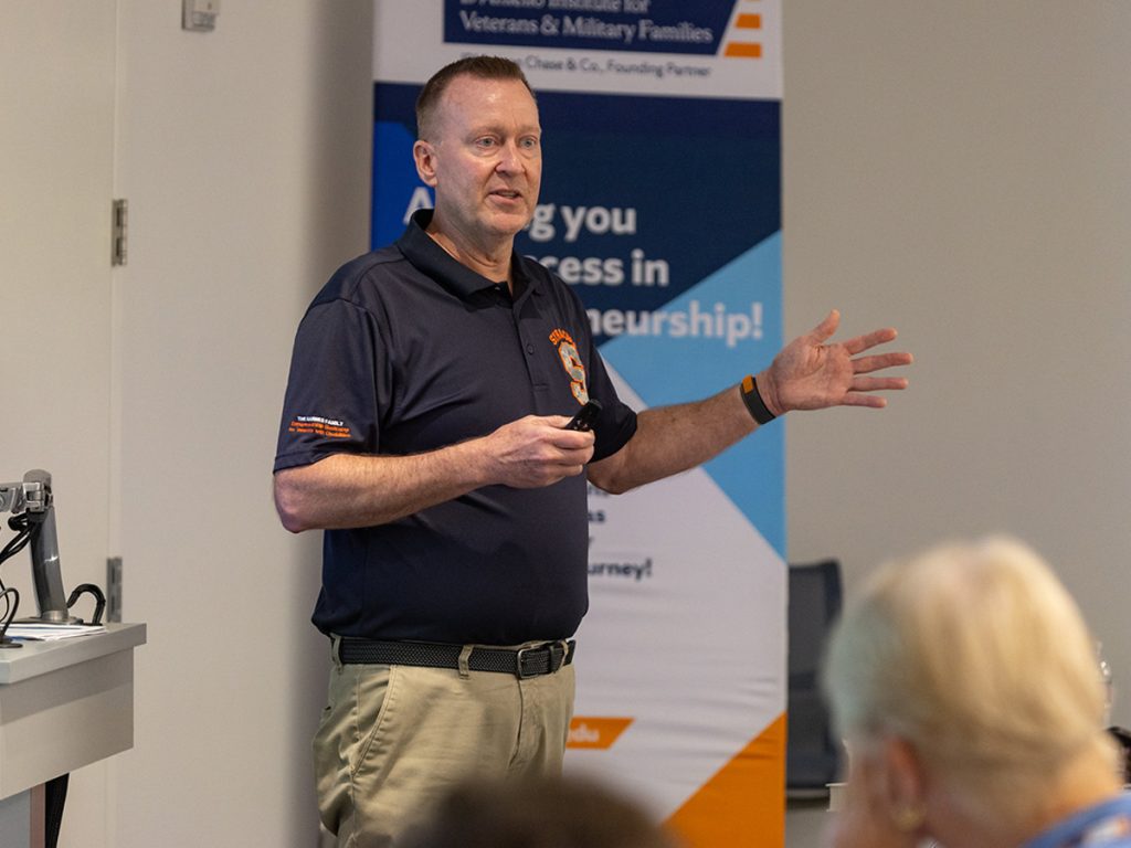 Mike Haynie speaks to an audience in front of a banner for the D'Aniello Institute for Veterans & Military Families entrepreneurship program