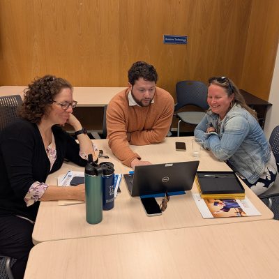 Three colleagues sit together at a conference table, leaning in to view a laptop screen. The room has wood-paneled walls and a small window.