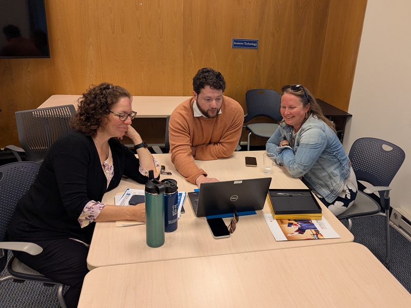Three colleagues sit together at a conference table, leaning in to view a laptop screen. The room has wood-paneled walls and a small window.