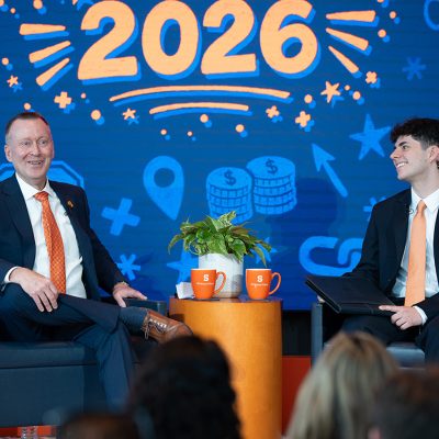 Two men in dark suits and orange ties sit on stage at a Syracuse University Whitman School event, with orange Whitman School mugs on a table between them and a '2026' graphic on the screen behind them.