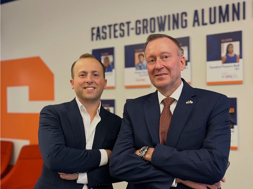 Two men in dark suits stand with arms crossed in front of a Syracuse University 'Fastest-Growing Alumni' wall display
