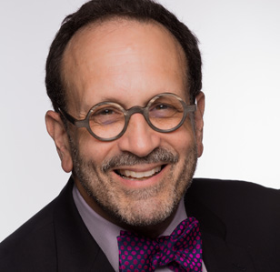 Headshot of a person wearing round glasses, a dark suit jacket, and a polka-dot bow tie, smiling against a plain light background.