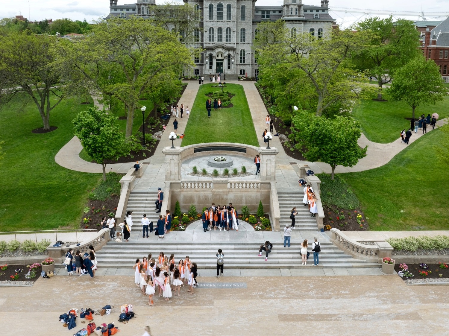 Overhead view of campus quad with steps, walkway, and groups of graduates in caps and gowns gathered around a central lawn and historic building.