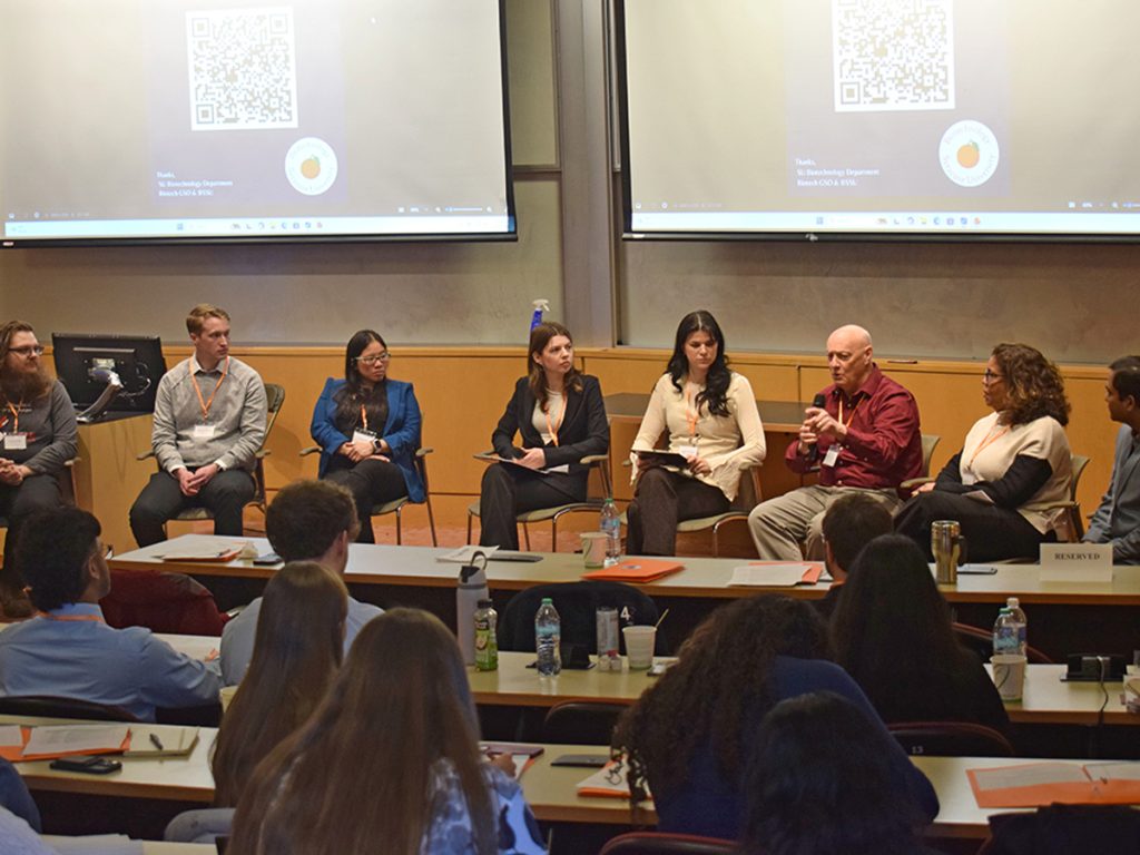 Eight panelists seated at the front of a lecture hall during a SU Biotechnology Department event, with an audience of students listening from tiered seating
