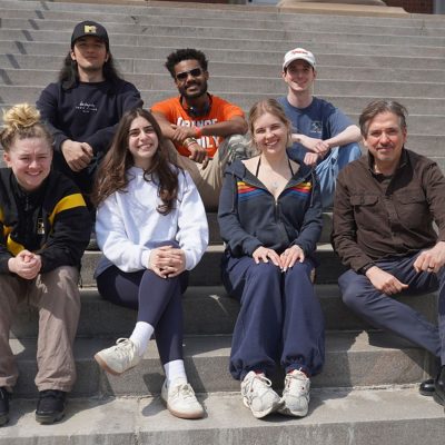 A group of seven students and a faculty member sit together on outdoor campus steps, smiling on a sunny day.
