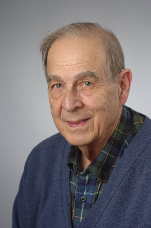 Studio portrait of an older adult wearing a blue cardigan over a plaid shirt, facing forward against a plain gray background.