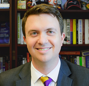 Professional headshot of a smiling man in a gray suit and gold-and-purple striped tie, with bookshelves in the background