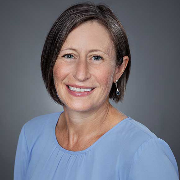 Headshot portrait of a person with short dark hair, wearing a light blue top and drop earrings against a gray background.