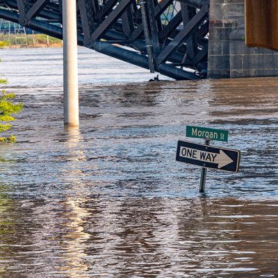 Floodwaters submerge Morgan Street near a large steel railroad or highway bridge, with a One Way sign partially underwater. Brown, murky water fills the street to several feet deep, with a light pole and green trees also visible in the background.
