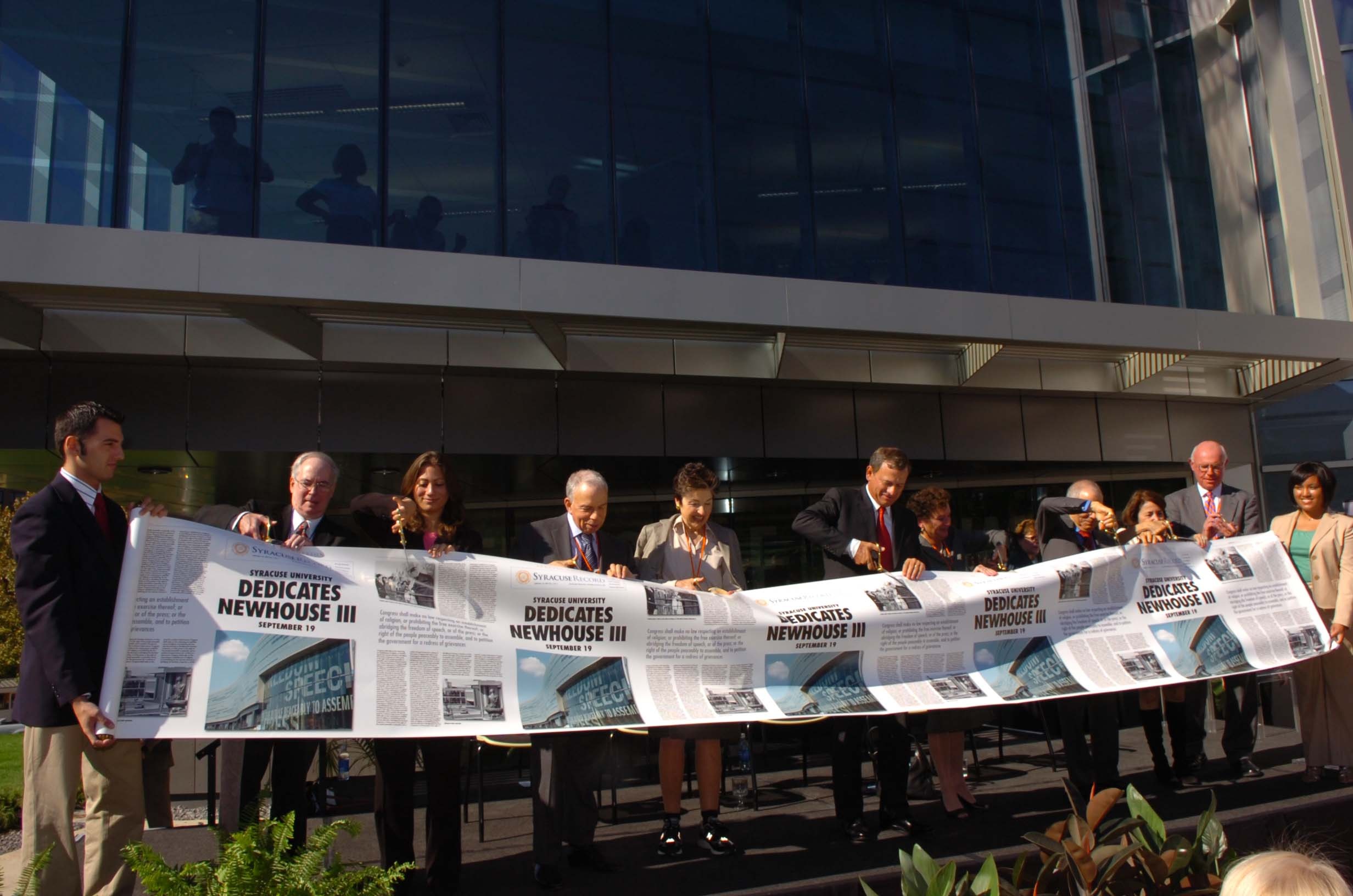 A line of dignitaries stands outside a modern glass building, holding a large banner designed to look like a newspaper with the headline "Dedicates Newhouse III." The group is participating in a ribbon-cutting-style ceremony for the Newhouse III building at Syracuse University.