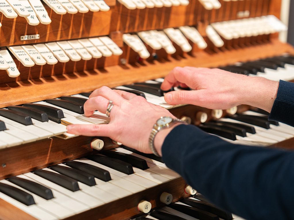 A close-up of hands playing a pipe organ console, with multiple keyboards and rows of labeled stop knobs on a warm wooden frame.
