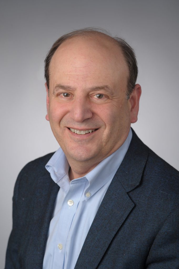 A professor smiles while posing for a headshot in front of a grey backdrop.