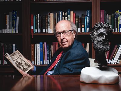 A man holds an open book at a desk beside a bronze bust, with a wall of bookshelves behind him.