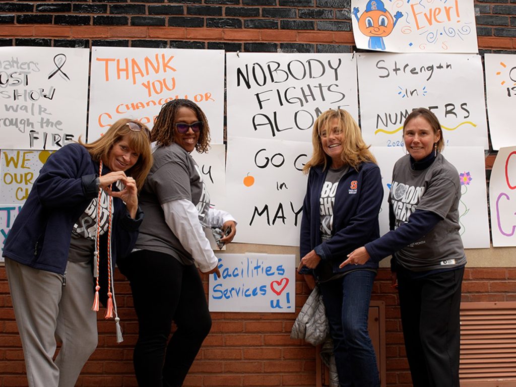 Four women pose and smile in front of a wall covered with handmade supportive signs, including "Nobody Fights Alone," "Thank You Chancellor," "Strength in Numbers," and "Facilities Services ♥ U!