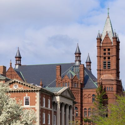 Historic red-brick academic buildings with ornate towers and a tall clock-like tower, framed by blooming white trees under a partly cloudy sky.