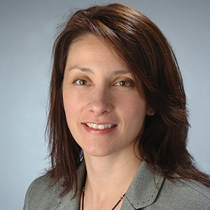 Professional headshot of a woman with shoulder-length auburn hair, wearing a gray blazer, smiling against a light blue background.