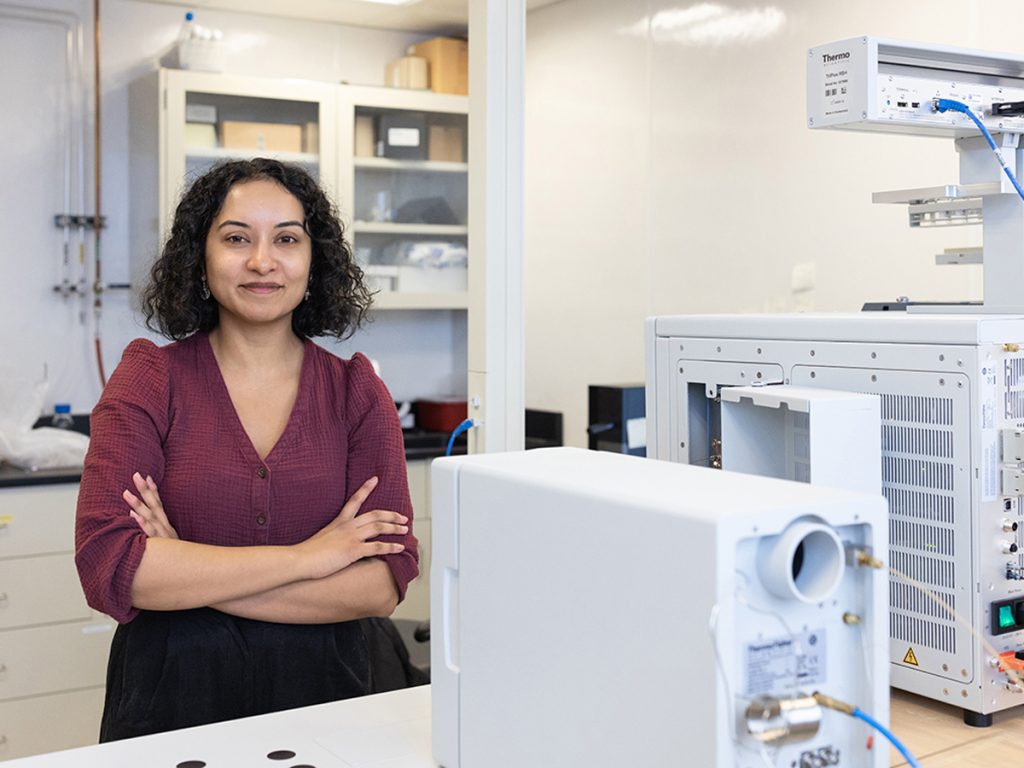 A woman with curly dark hair stands with arms crossed and smiling in a scientific laboratory, surrounded by large white Thermo Scientific instruments and equipment, with storage cabinets visible in the background.