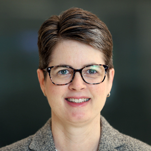 A person smiles for a headshot wearing glasses and a gray blazer.