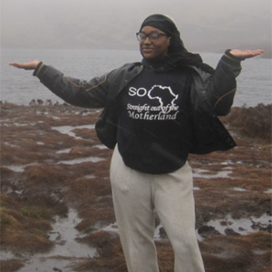 A person standing outdoors with arms outstretched, wearing a black jacket that reads "Straight out of the Motherland," with a gray coastal landscape in the background