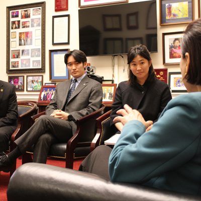 Three young professionals seated in an office meeting with a government official, surrounded by framed photos and awards on the wall."