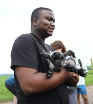 A young man in a black t-shirt holds two small baby goats outdoors on a farm.