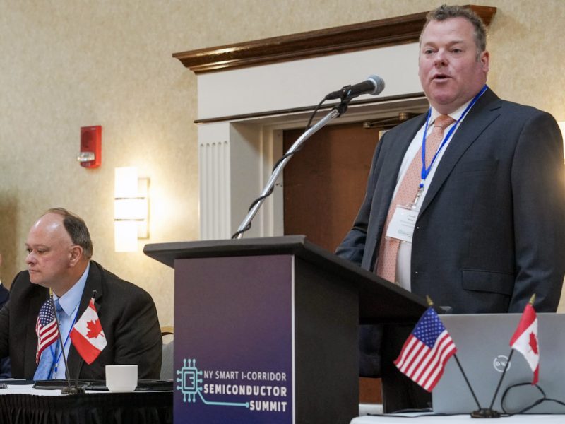 Panelists seated beside a speaker at a podium during the NY SMART I-Corridor Semiconductor Summit, with U.S. and Canadian flags displayed.