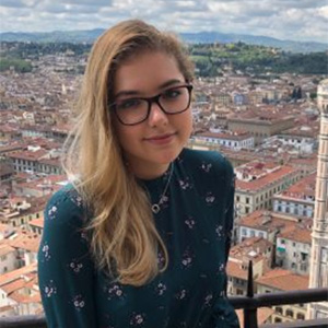 A young woman with long blonde hair and glasses smiling in front of a panoramic view of a European city.