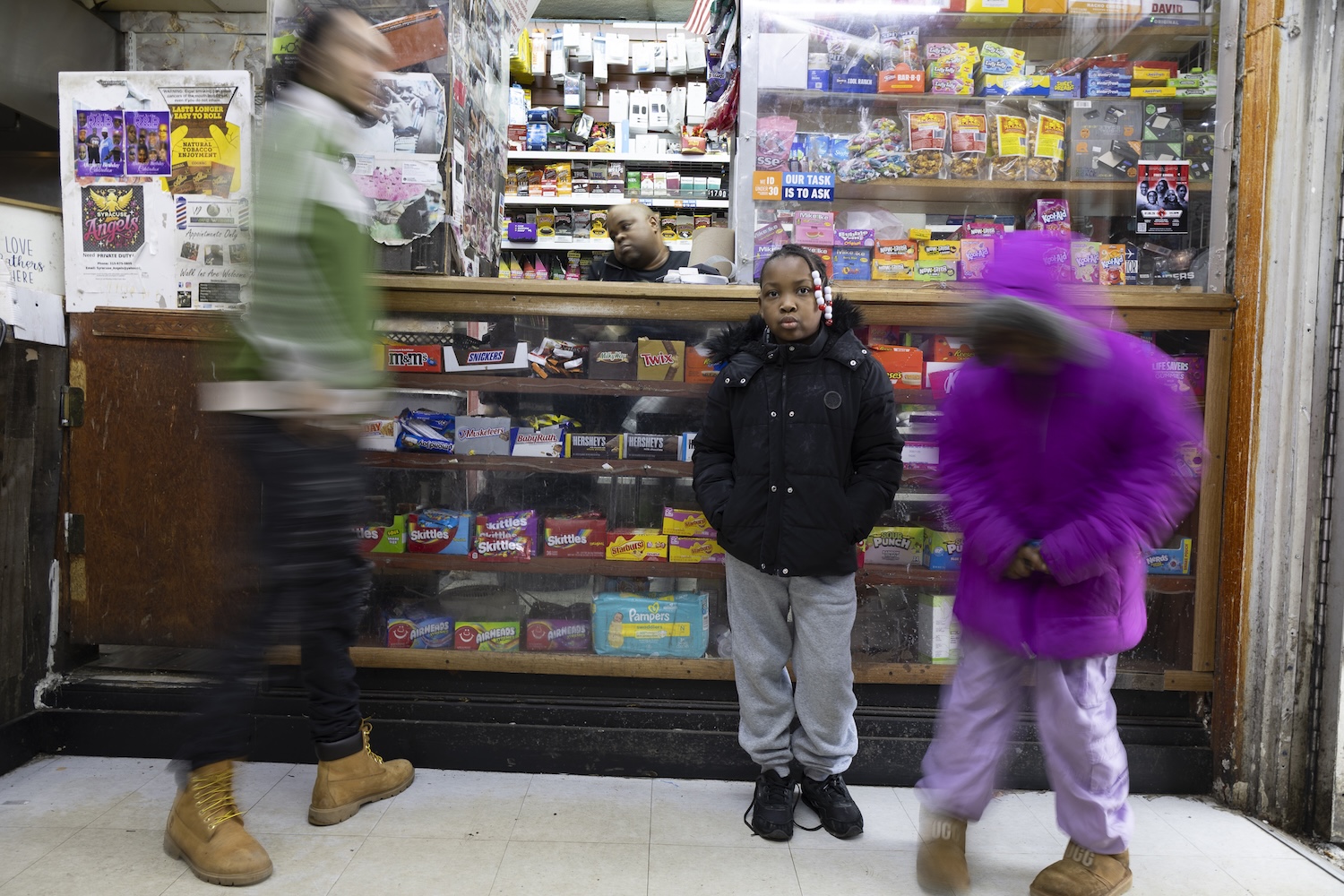 Child standing still in front of a store counter filled with candy, while two other figures pass by in motion blur.