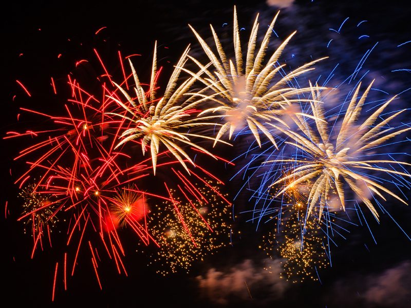 Multiple fireworks bursting in a dark night sky, with red starbursts on the left, golden chrysanthemum-shaped explosions in the center, and blue streaks on the right, with falling golden sparks below.