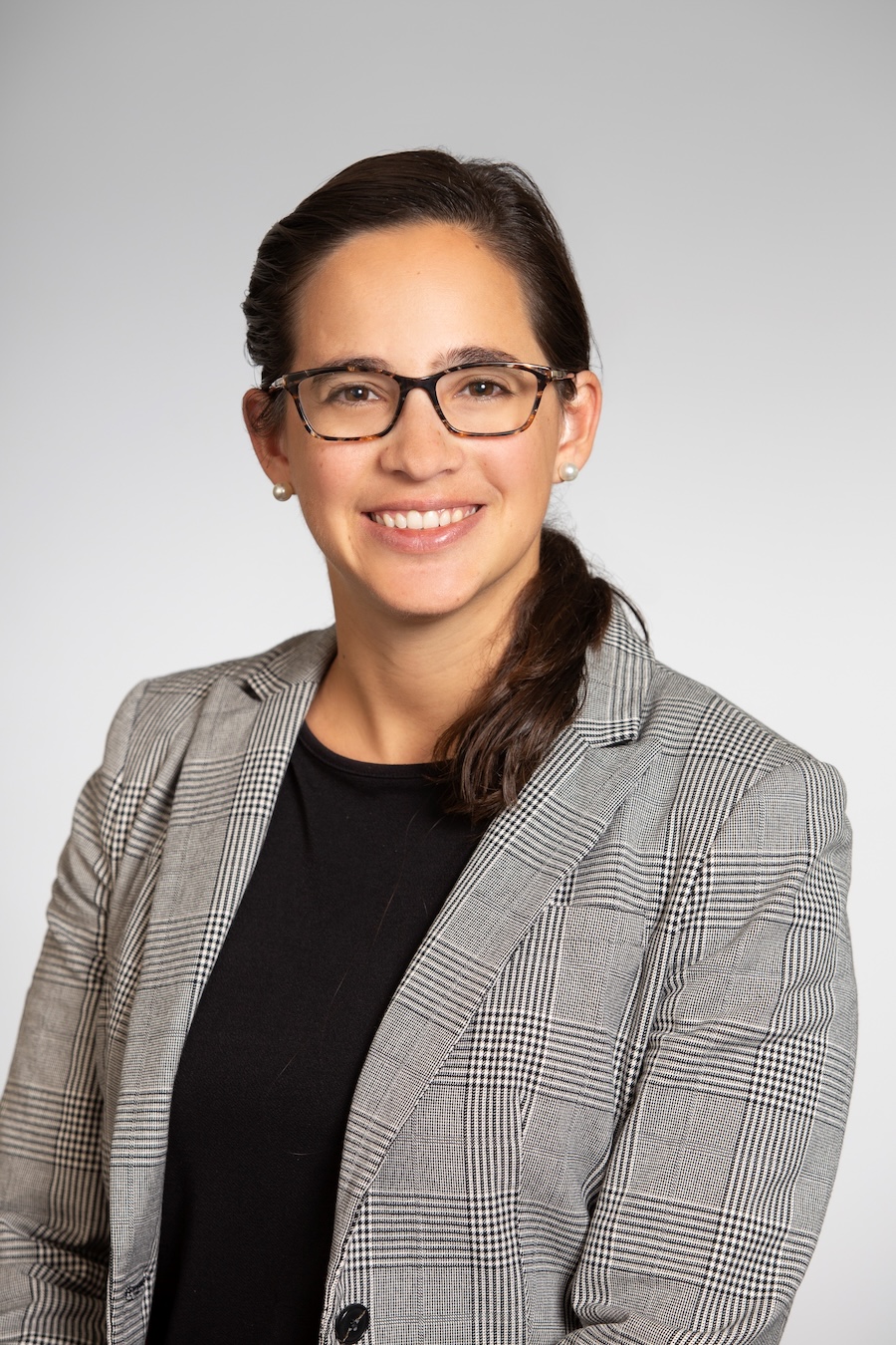 Headshot of a person wearing glasses, a gray plaid blazer, and a black top, smiling against a plain light background.