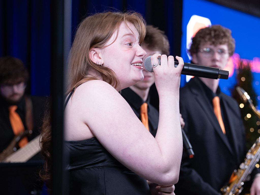 A female vocalist performs at an indoor event, holding a microphone and smiling mid-song, with jazz ensemble members dressed in black with orange ties visible in the background