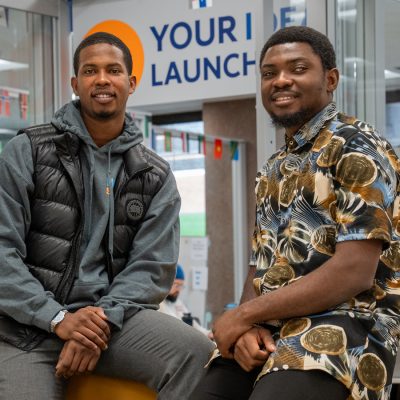 Two male students sit and smile in front of a "Your Idea Launches Here" sign at LaunchPad.
