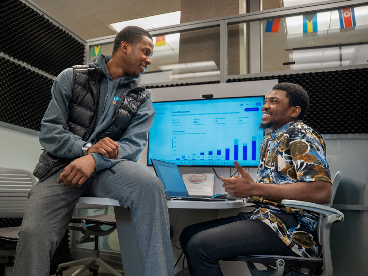 Two male students laugh together at a table displaying the GritGateway platform on a large monitor.