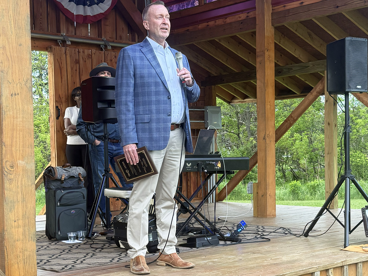 A man in a blue blazer speaks into a microphone on an outdoor pavilion stage while holding a WCNY Honor Our Heroes award plaque.