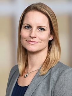 A woman with blond shoulder length hair is wearing a gray blazer and navy top, posed against a softly lit indoor background.