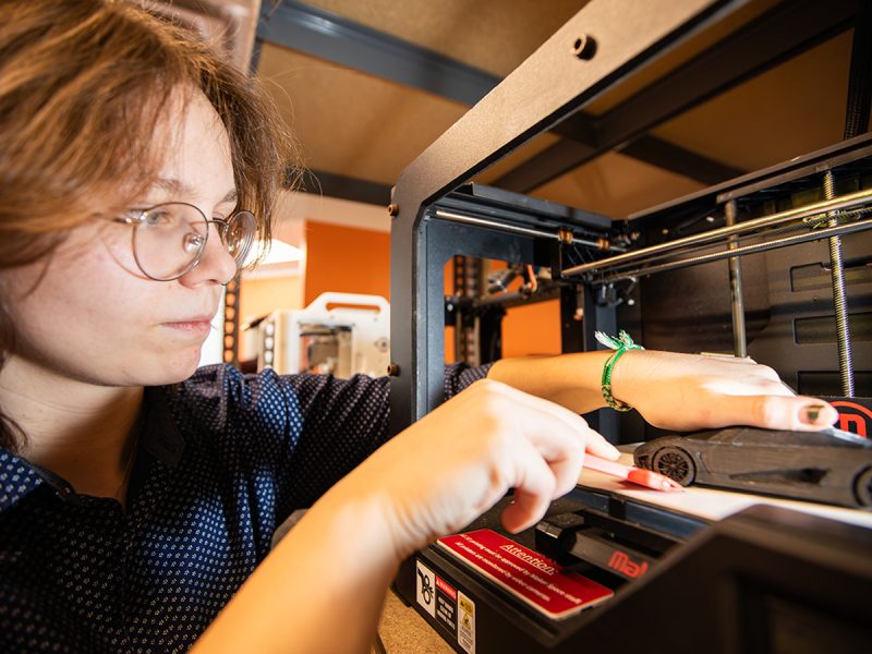 A person with glasses and curly hair examines a 3D printer, removing a small printed object from the print bed.