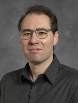 A person with short dark hair wearing glasses and a dark button-down shirt poses against a gray studio background.