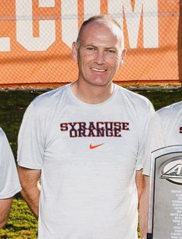 Man wearing a Syracuse Orange Nike shirt smiles outdoors near a soccer field