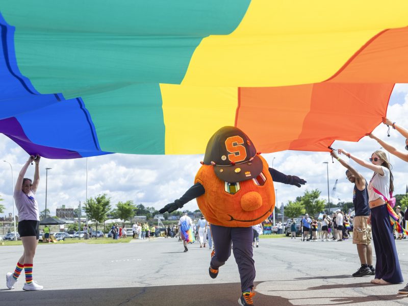 Otto the Orange dances under a large rainbow Pride flag at a parade.